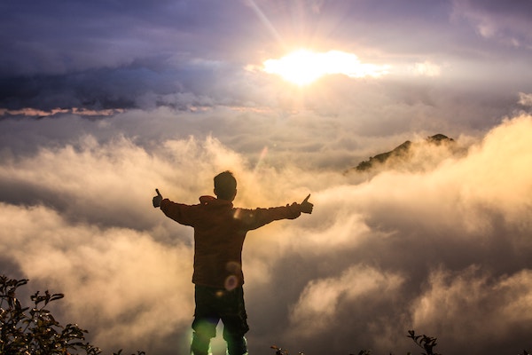 Man standing on mountain top stretching out his arms looking at clouds and sun, ari monkarsh achieve success