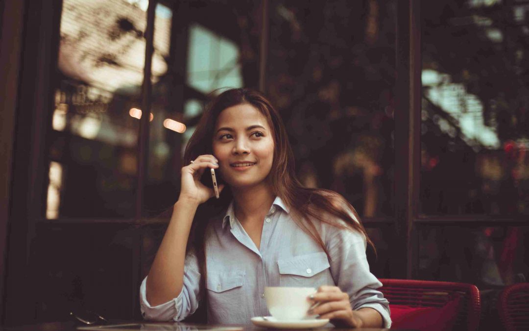 Woman sitting at a table outside of a cafe holding a phone and smiling while glancing to the side, image used for Ari Monkarsh blog on professionals lack the skill of communication