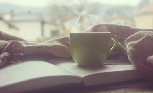 Cup of coffee sitting on top of an open book with a blanket and window in the background, image used for Ari Monkarsh blog on creating a successful morning routine